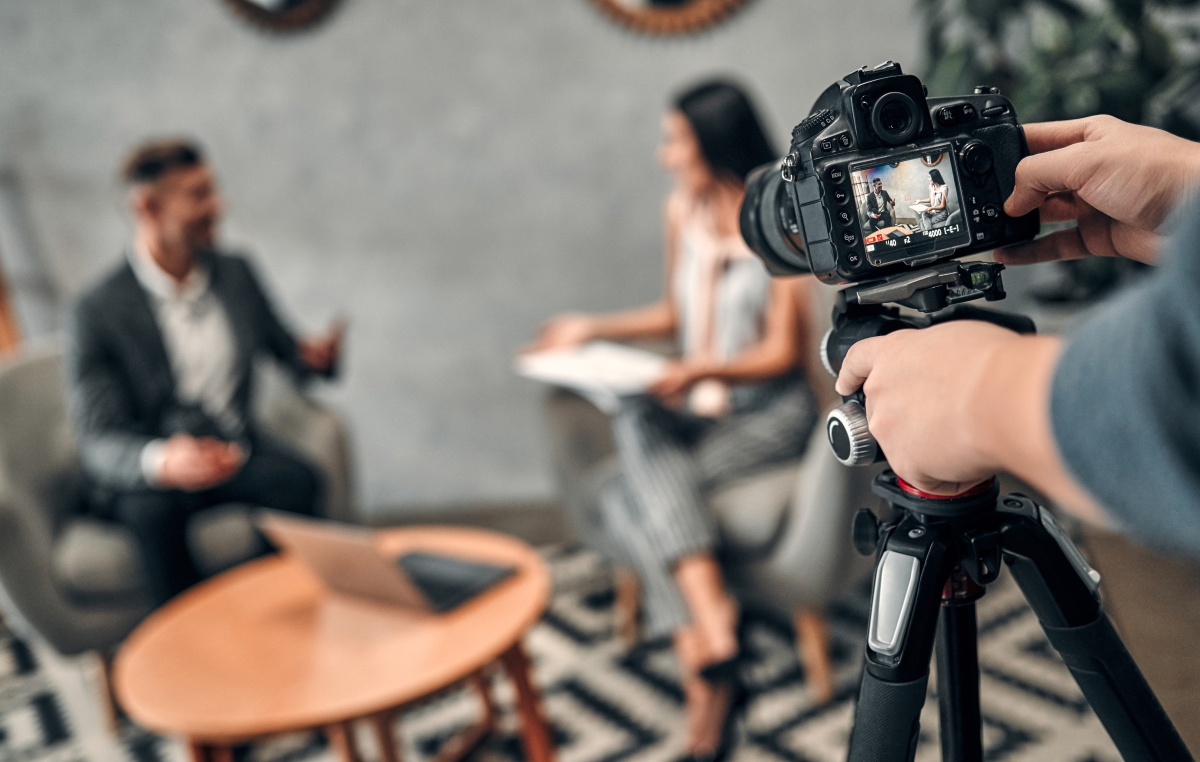 Im Vordergrund ein Stativ mit Kamera, im Hintergrund findet ein Interview statt, unscharf zu sehen sind eine weibliche und eine männliche Person in einer Interviewsituation.